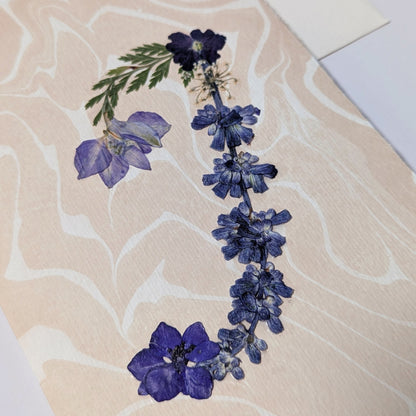 Handcrafted pressed flower initial “J” on  blush neutral Suminagashi marbled paper, featuring deep blue and violet florals with delicate green foliage.