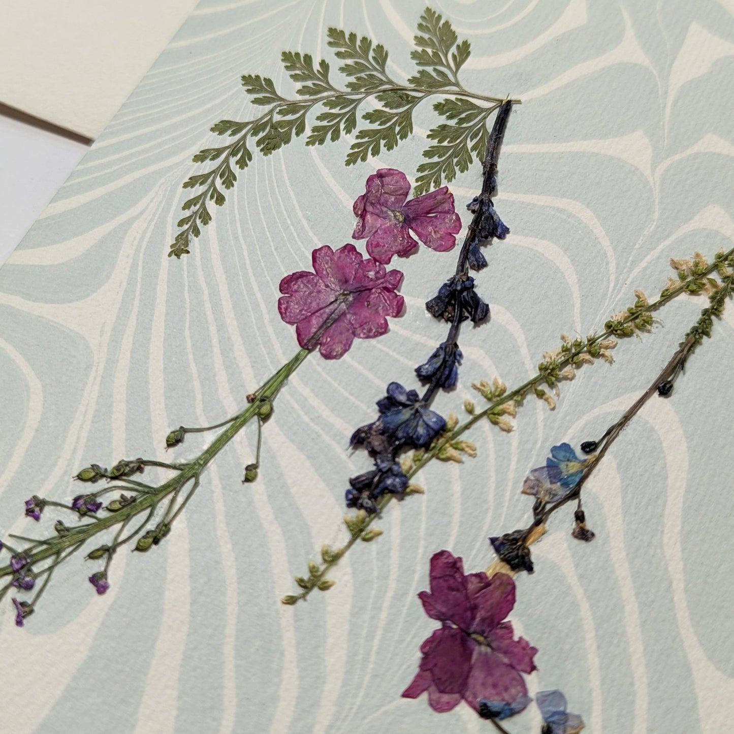 Pressed flower “M” initial notecard with pink and blue botanicals on marbled Suminagashi background, handmade on Fabriano Medioevalis paper