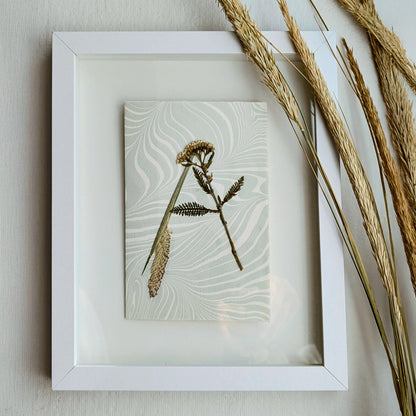 Framed botanical initial "A" artwork on Suminagashi marbled paper displayed with dried grasses, showcasing handmade pressed floral notecard