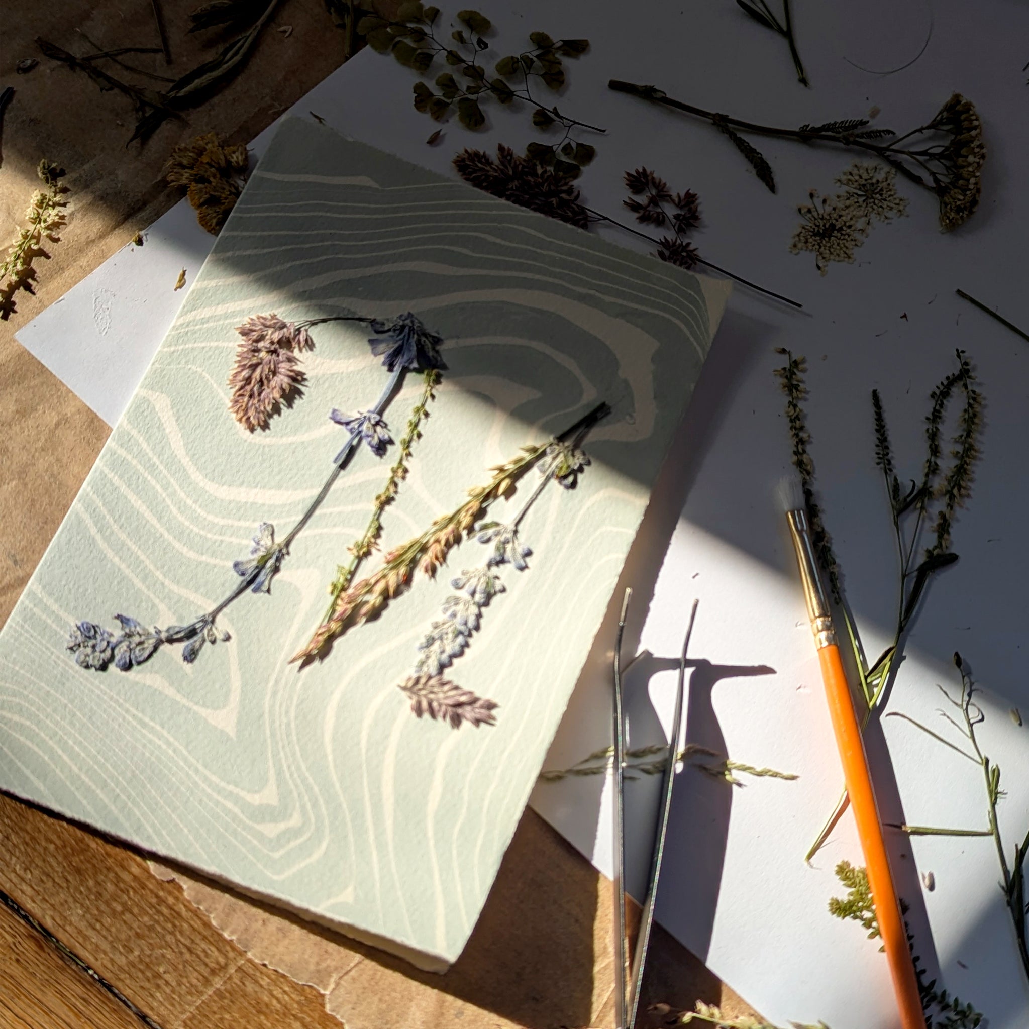 Pressed flower initial notecard in progress with tools and botanicals, showcasing handmade Suminagashi paper and artisan process