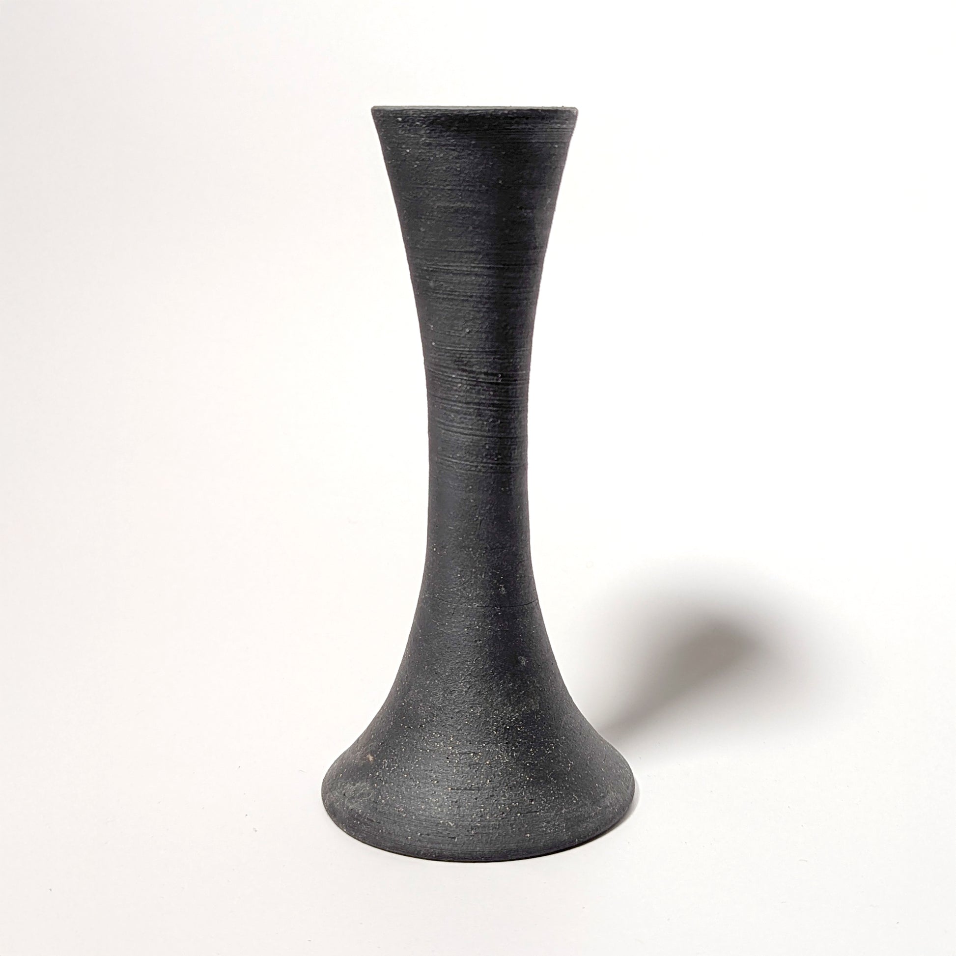 A single handcrafted black clay candle taper holder stands against a white backdrop. The wheel-thrown form shows subtle ridges and a matte, stone-like texture, highlighting its modern organic silhouette.