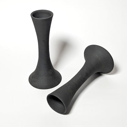 Two handcrafted black clay candle taper holders displayed on a white background. One stands upright while the other lies horizontally, revealing smooth throwing rings and organic curves.