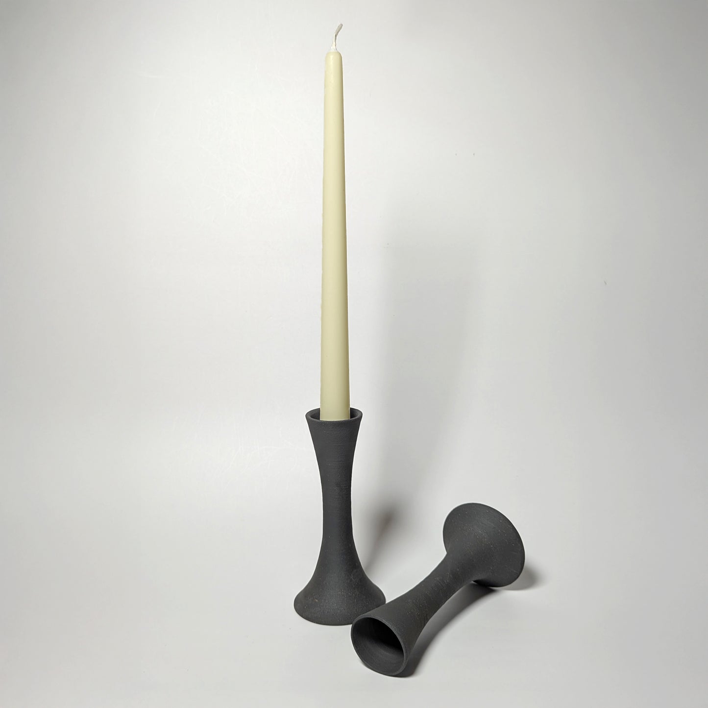 Two black clay candle taper holders on a white surface—one upright holding a cream taper candle, the other resting on its side—showing elegant, sculptural contours and a softly textured matte finish.
