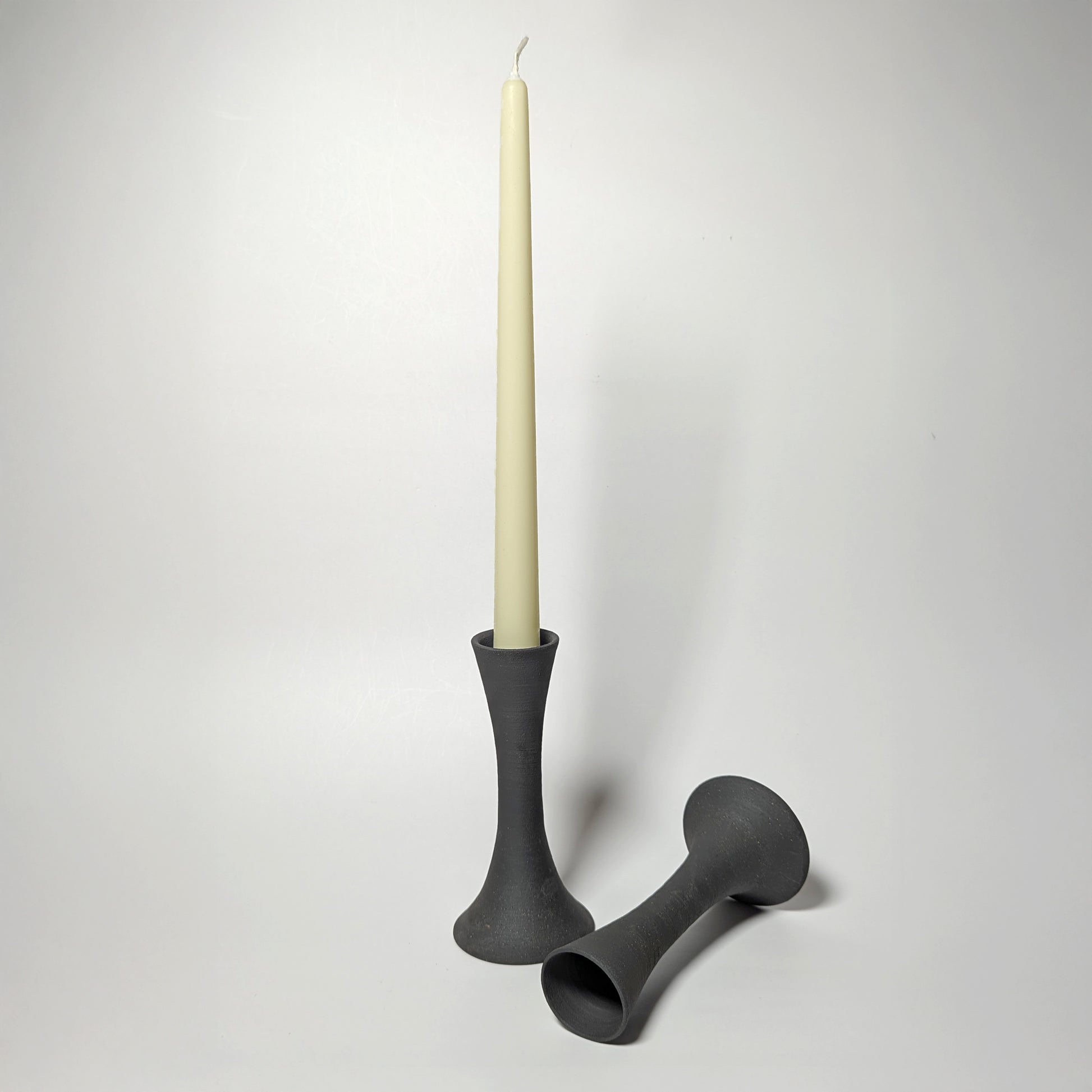 Two black clay candle taper holders on a white surface—one upright holding a cream taper candle, the other resting on its side—showing elegant, sculptural contours and a softly textured matte finish.