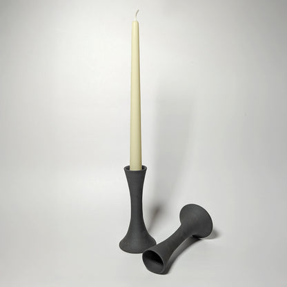 Two black clay candle taper holders on a white surface—one upright holding a cream taper candle, the other resting on its side—showing elegant, sculptural contours and a softly textured matte finish.