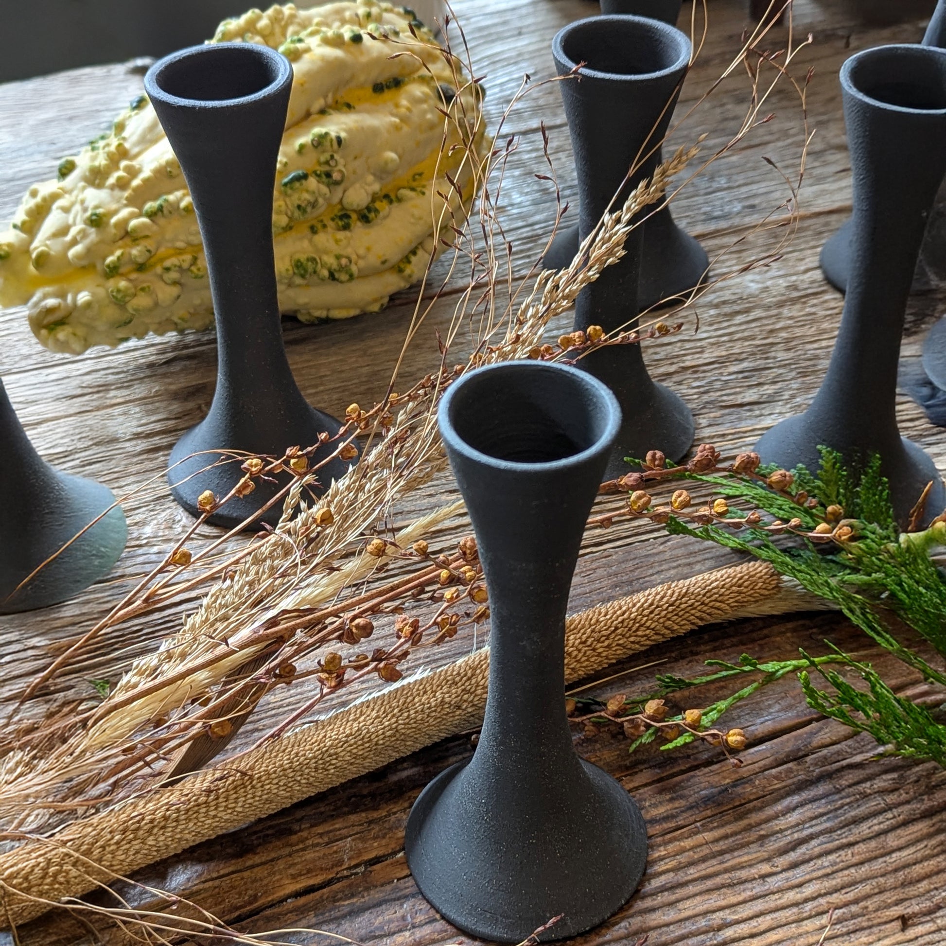 Close-up of multiple black clay candle holders among dried grasses, green cedar sprigs, and a textured gourd, highlighting the earthy matte tones and handcrafted form.