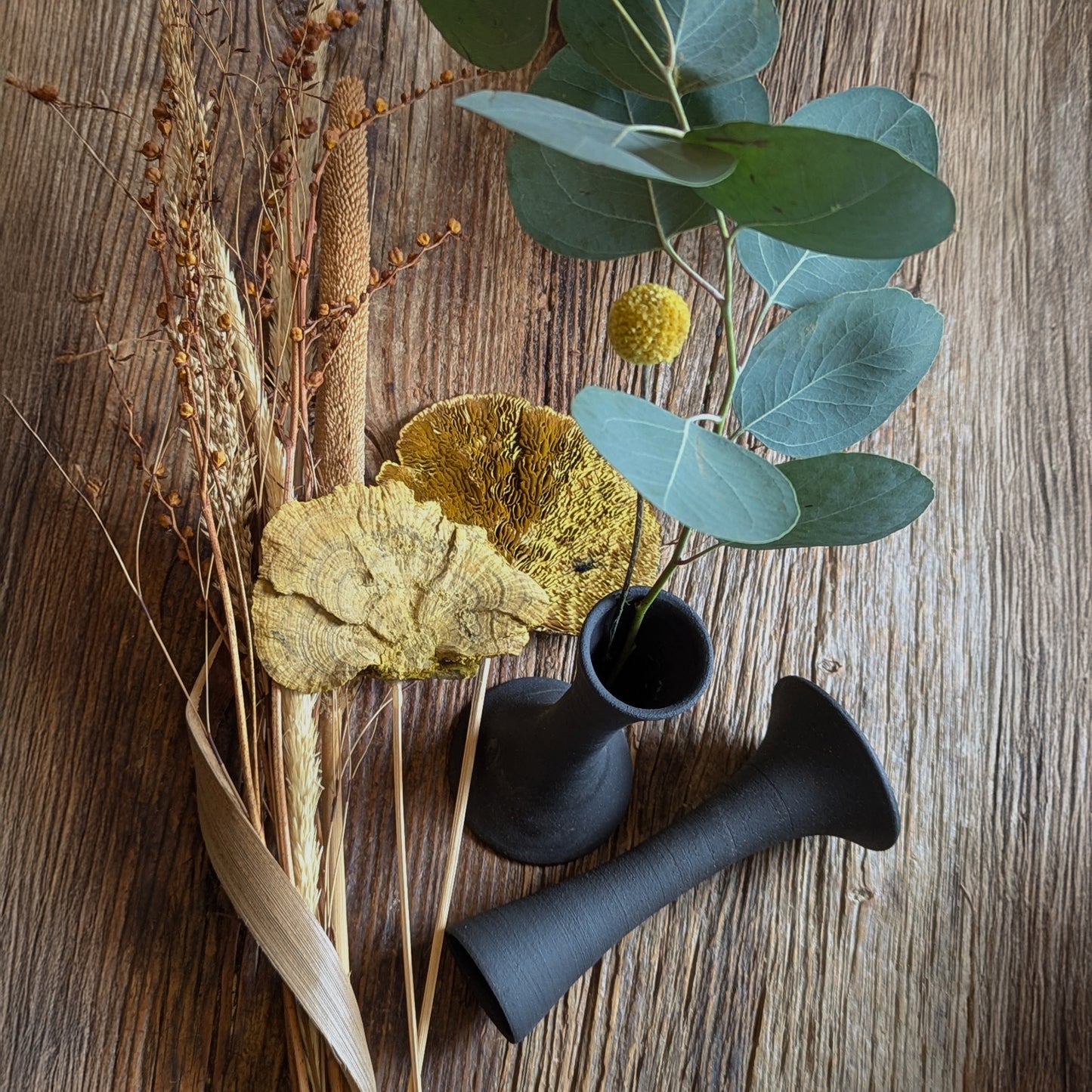 Overhead view of two black clay taper holders styled with dried florals, eucalyptus, and sculptural mushrooms on a wooden surface, showcasing their versatility as dry stem vessels.