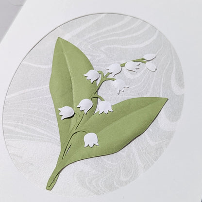 Lily of the valley paper cut floral design on hand-marbled suminagashi background with soft green leaves and white blossoms