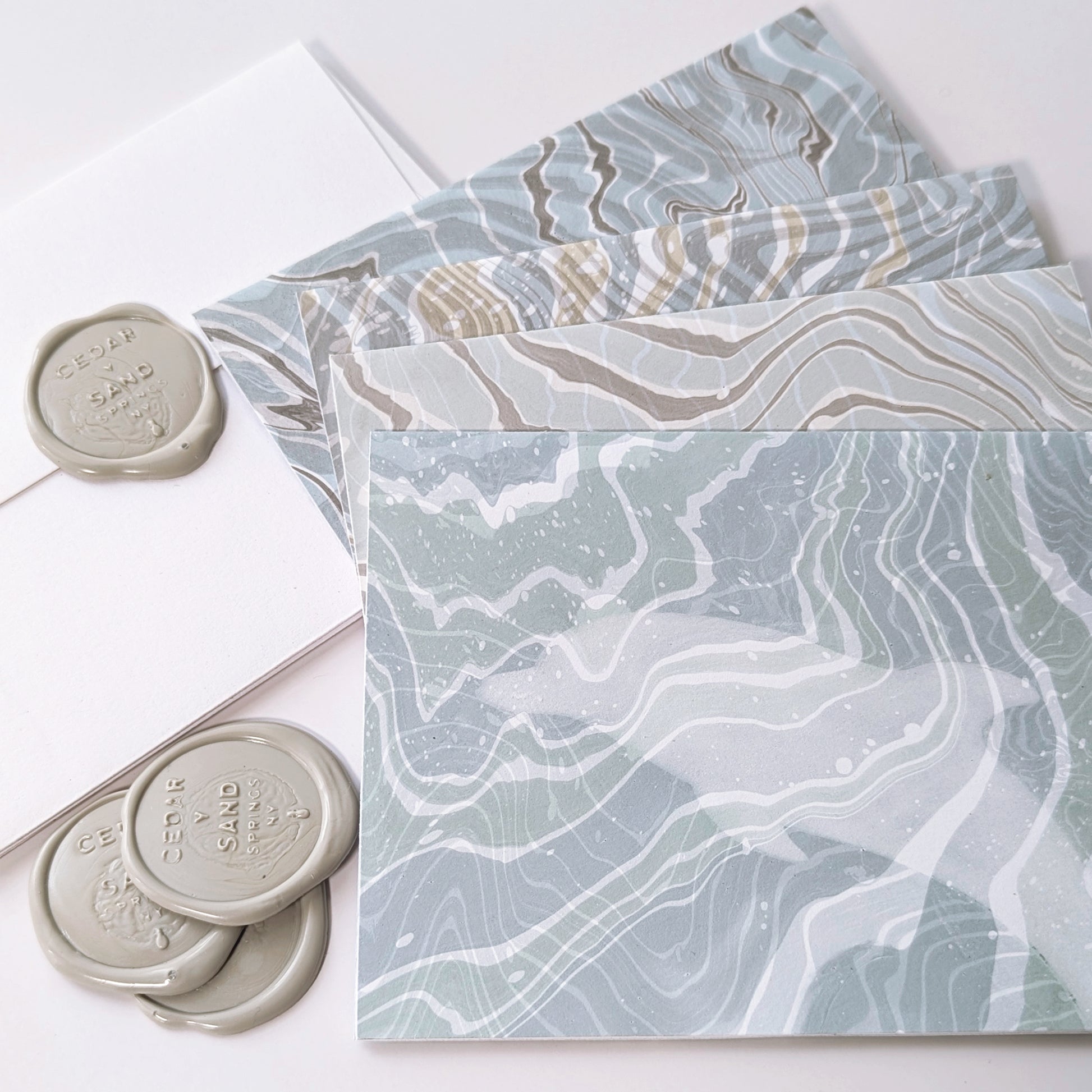Detail of a Marine Fauna Fin Series notecard showing a dolphin silhouette masked into swirling blue, white, and gray suminagashi marbling with gray Cedar Y Sand insignia self-adhesive wax seals.