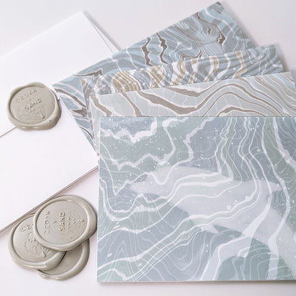 Detail of a Marine Fauna Fin Series notecard showing a dolphin silhouette masked into swirling blue, white, and gray suminagashi marbling with gray Cedar Y Sand insignia self-adhesive wax seals.