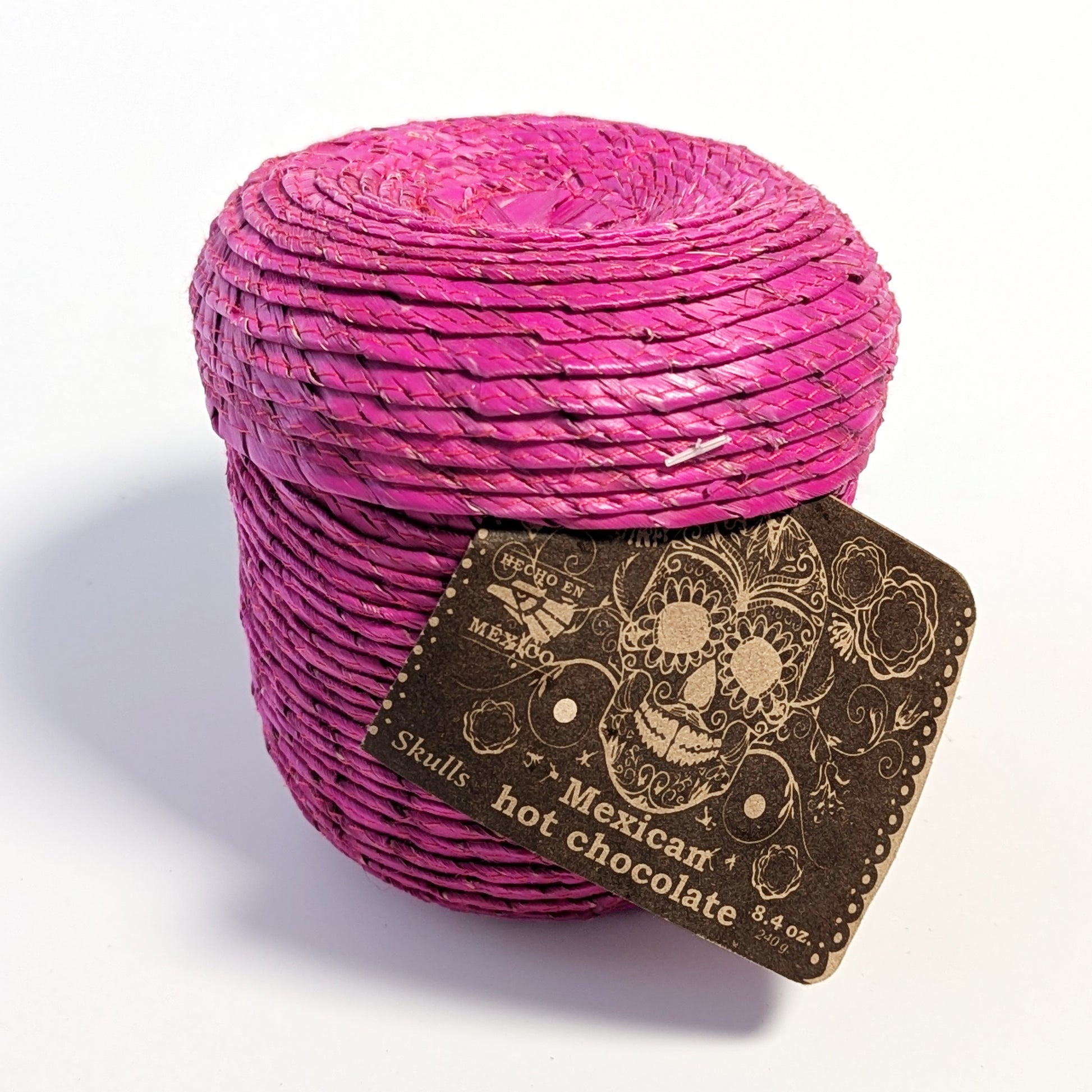 Lidded magenta palm leaf woven basket with dark brown label with skull illustration on a white surface.