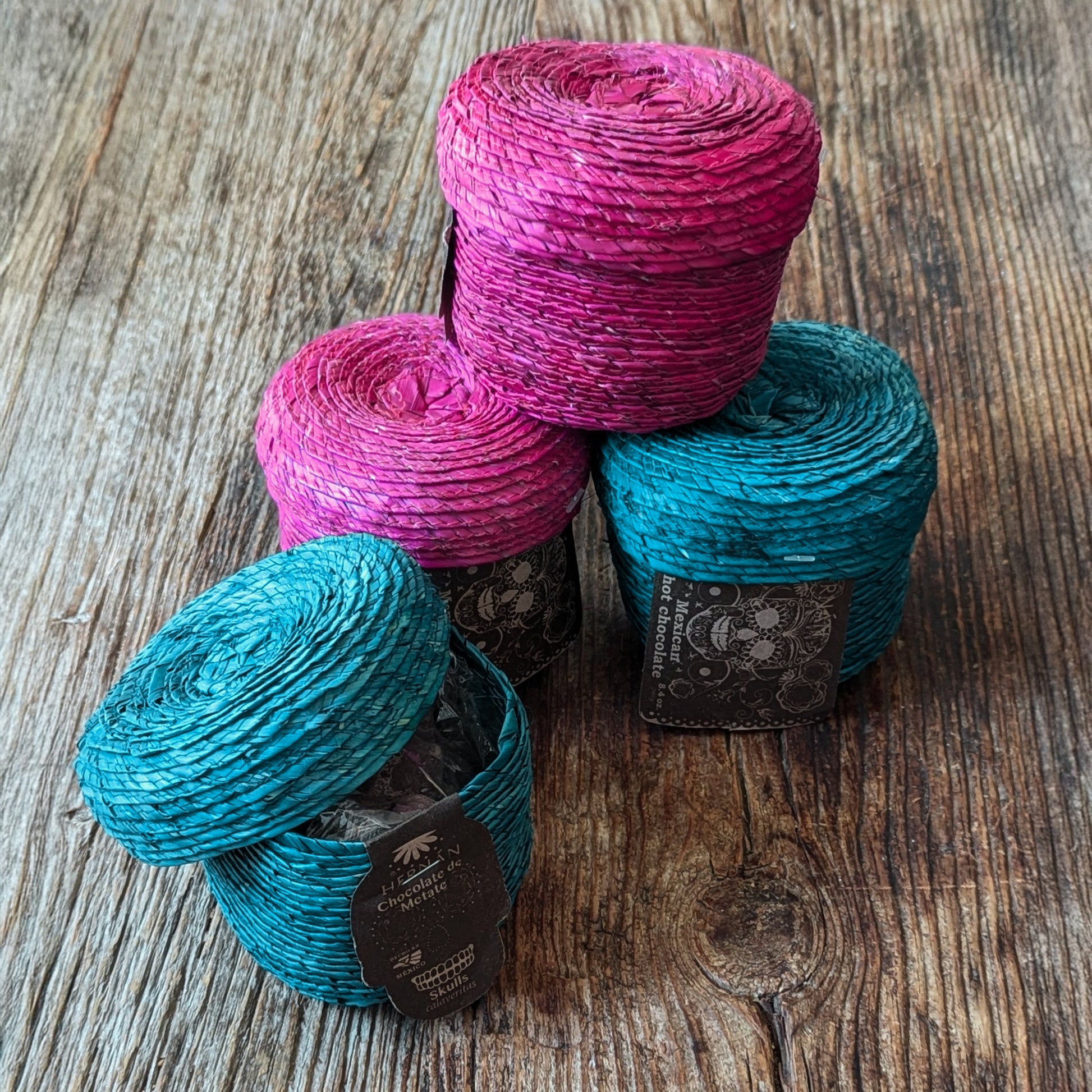 stacked image of 4 palm baskets, two teal and two magenta, each filled with handmade Mexican chocolate skulls, displayed on a rustic wooden surface.