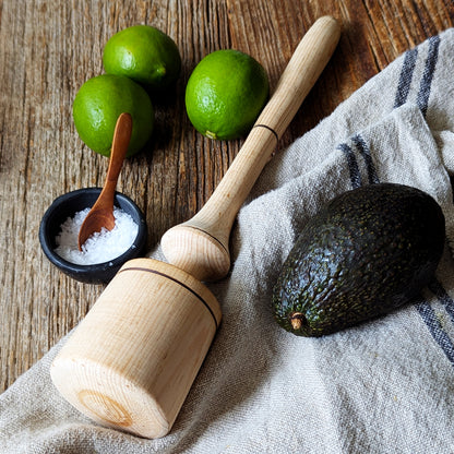 Wooden muddler, avocado, limes and sea salt on a rustic wooden countertop with a linen kitchen towel.