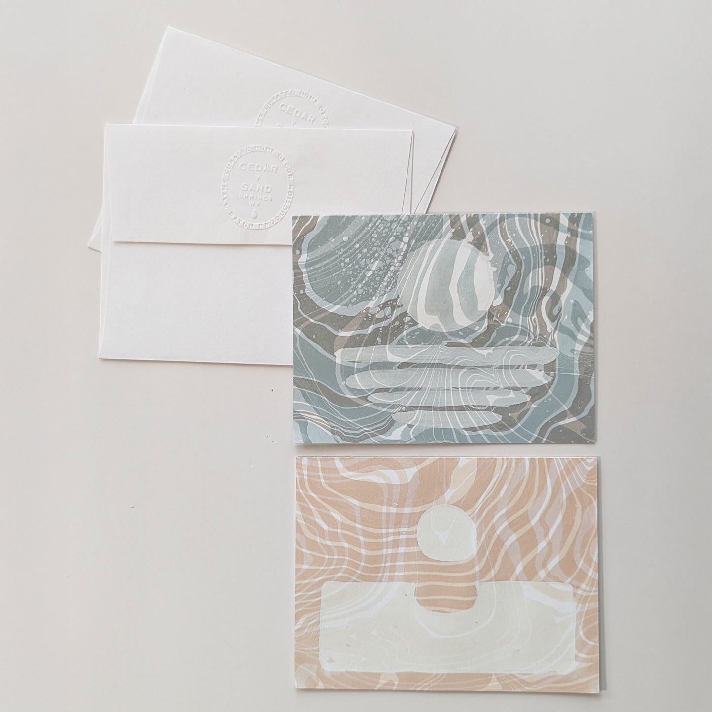 two hand-marbled Suminagashi notecards, one in dawn blue with a coastal moonset motif and one in coral with a sunrise design, displayed with matching envelopes.