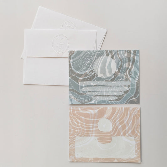 two hand-marbled Suminagashi notecards, one in dawn blue with a coastal moonset motif and one in coral with a sunrise design, displayed with matching envelopes.