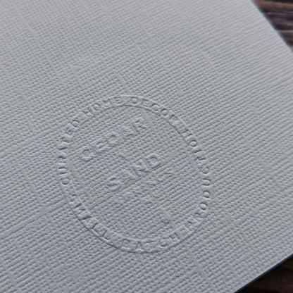 Textured warm gray Wheat Suminagashi notecard showing the embossed CEDAR Y SAND studio insignia on the reverse.