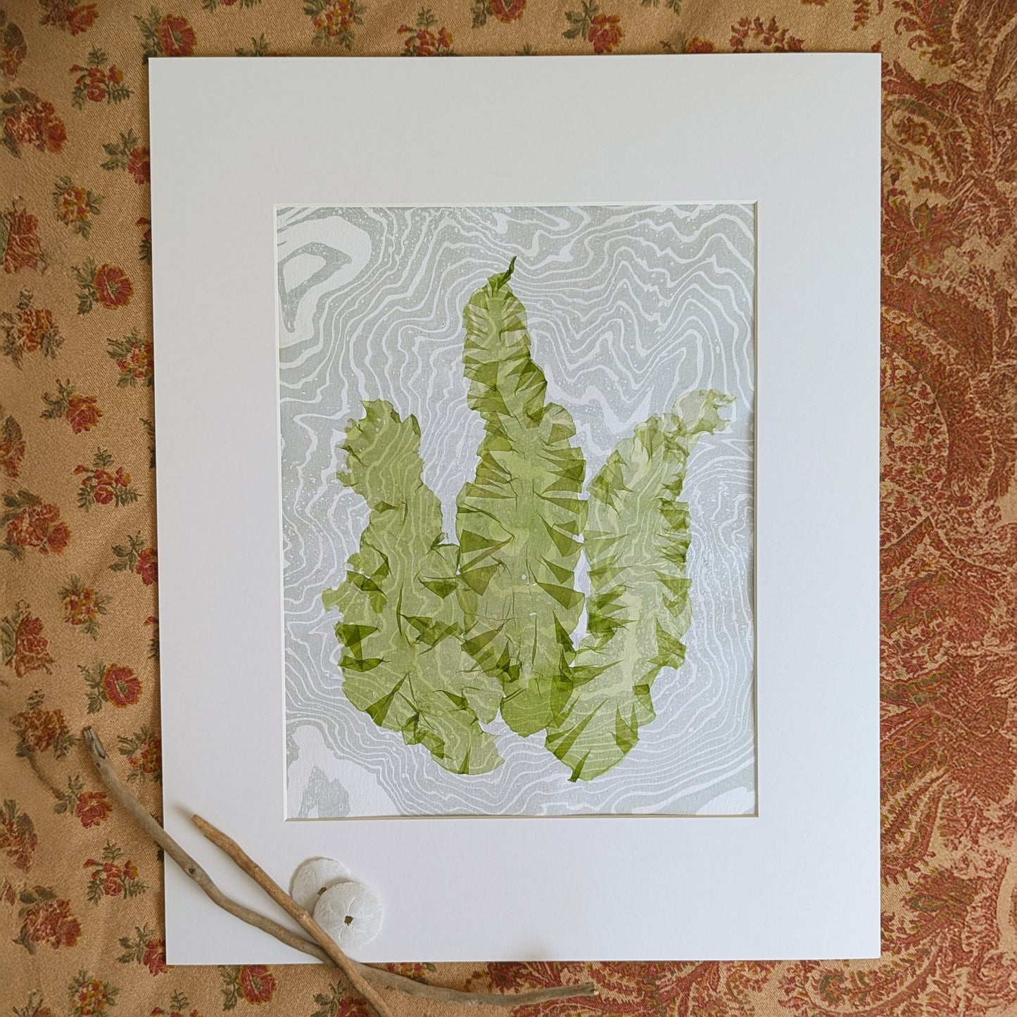 Framed pressed seaweed artwork with green Ulva lactuca on marbled paper, styled on patterned textile with driftwood accents