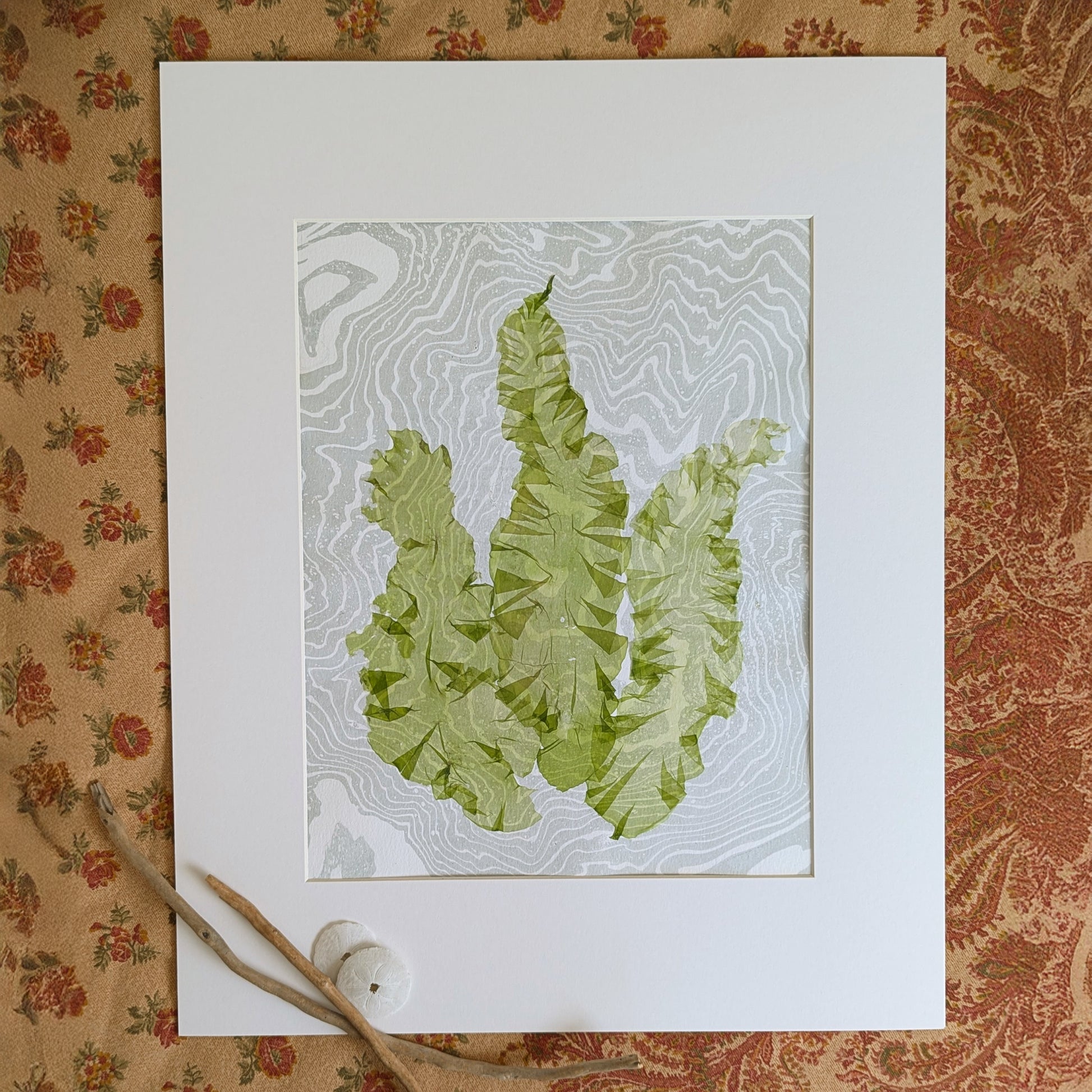 Framed pressed seaweed artwork with green Ulva lactuca on marbled paper, styled on patterned textile with driftwood accents
