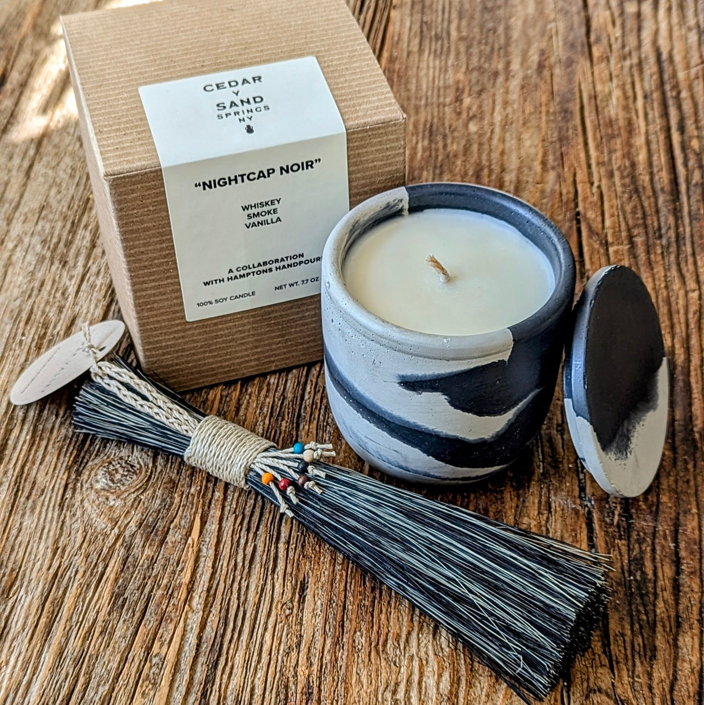 Artisan Hosting Set displayed on a rustic wood surface, featuring a Nightcap Noir soy candle in a smoke-fired ceramic vessel with its matching lid beside a kraft box, paired with a hand-wrapped Mixed Agave Slim Table Broom adorned with colorful thread accents.