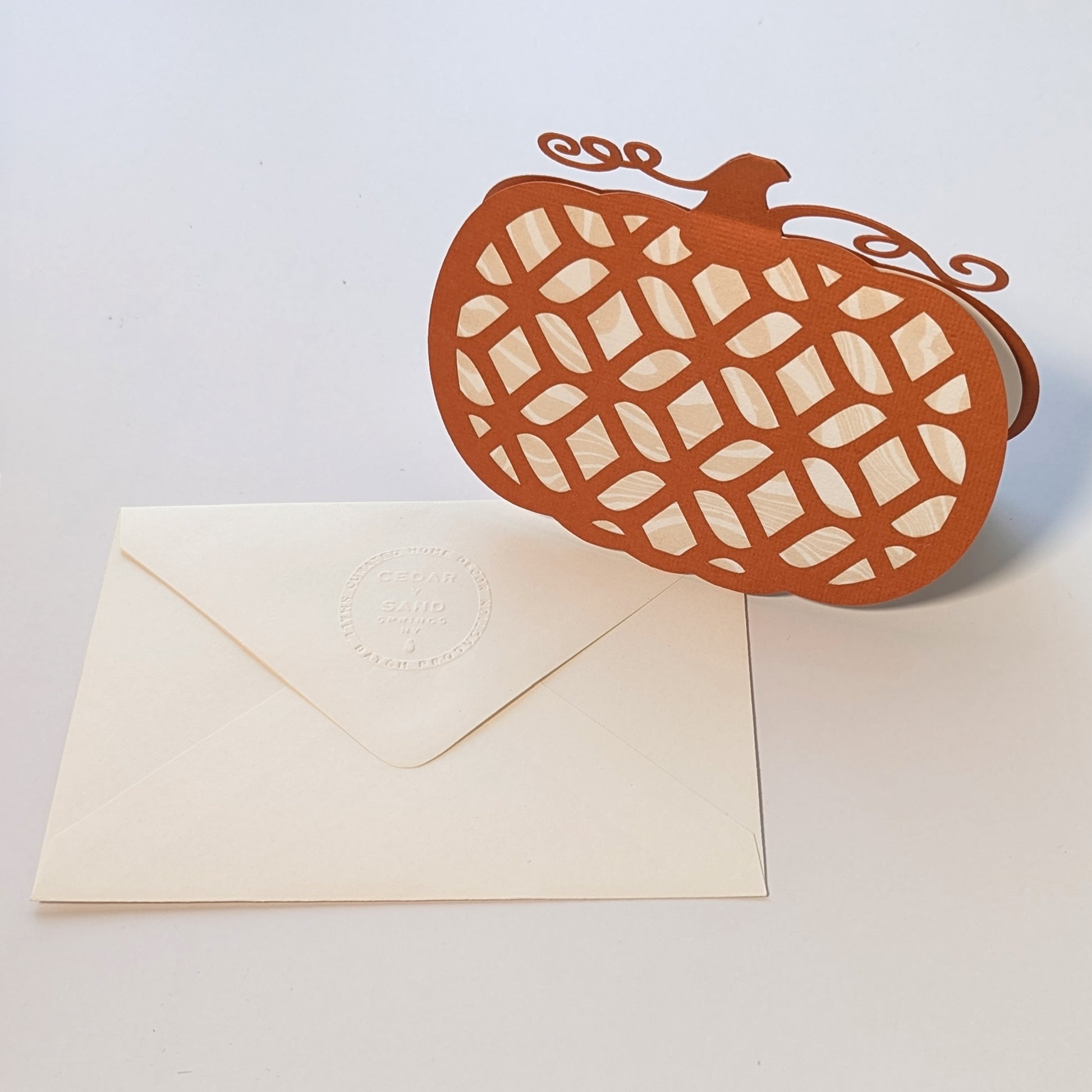 Front view standing beside embossed envelope on white background.
Alt: Pumpkin Lattice Notecard with cream envelope featuring CEDAR Y SAND embossed insignia.