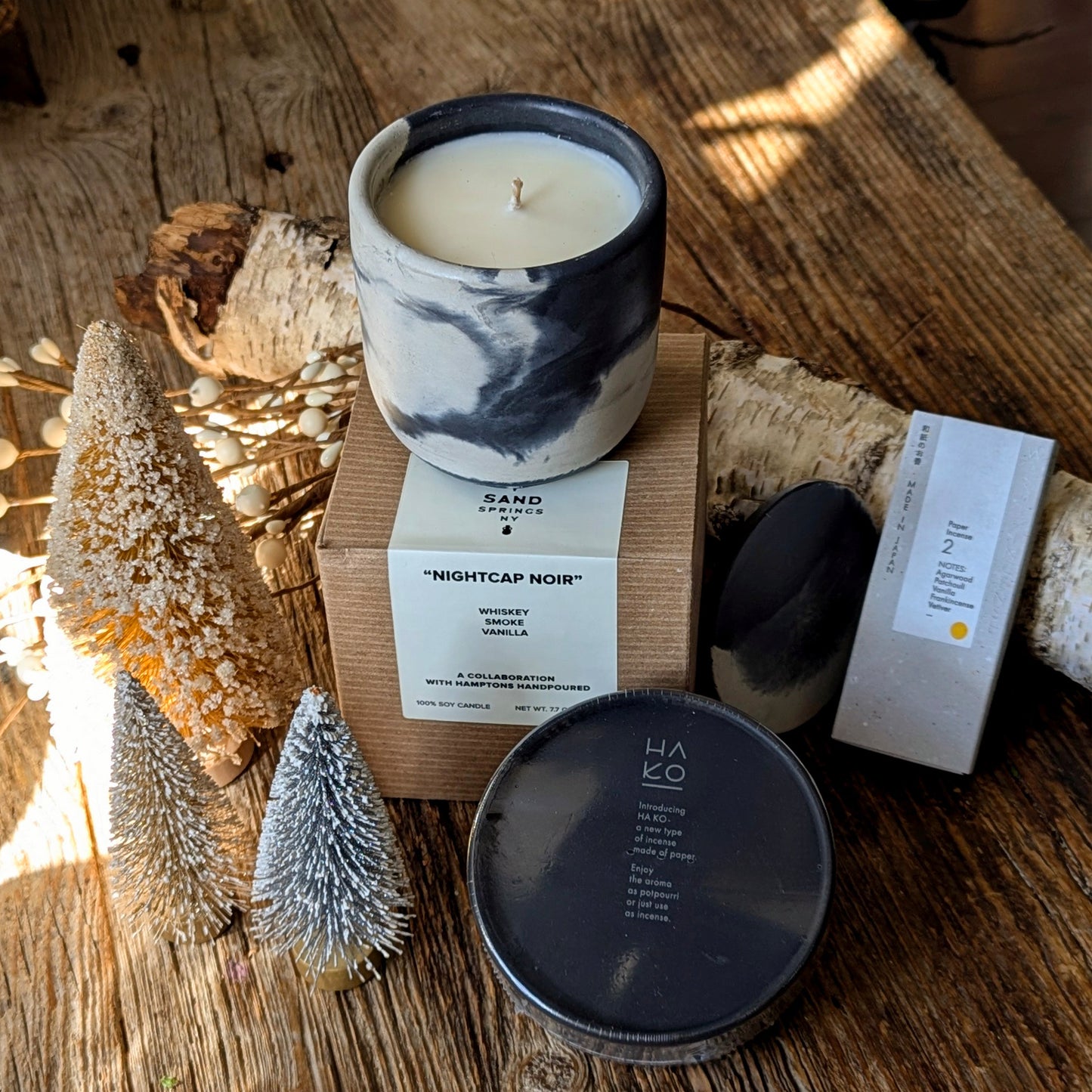Nightcap Noir candle in a black-and-white marbled vessel sits on its box beside HA KO black incense leaf packaging and Kunjudo agarwood washi incense strips, styled on a rustic wood surface with small winter bottlebrush trees and birch branches.