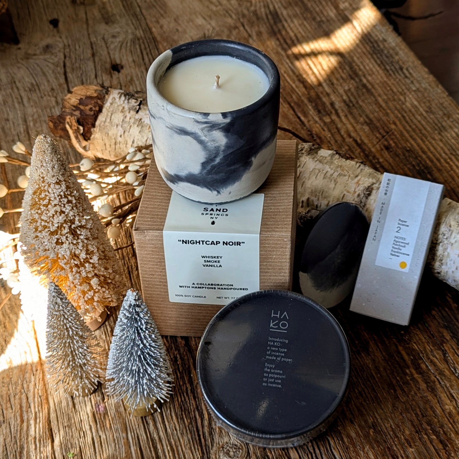 Nightcap Noir candle in a black-and-white marbled vessel sits on its box beside HA KO black incense leaf packaging and Kunjudo agarwood washi incense strips, styled on a rustic wood surface with small winter bottlebrush trees and birch branches.