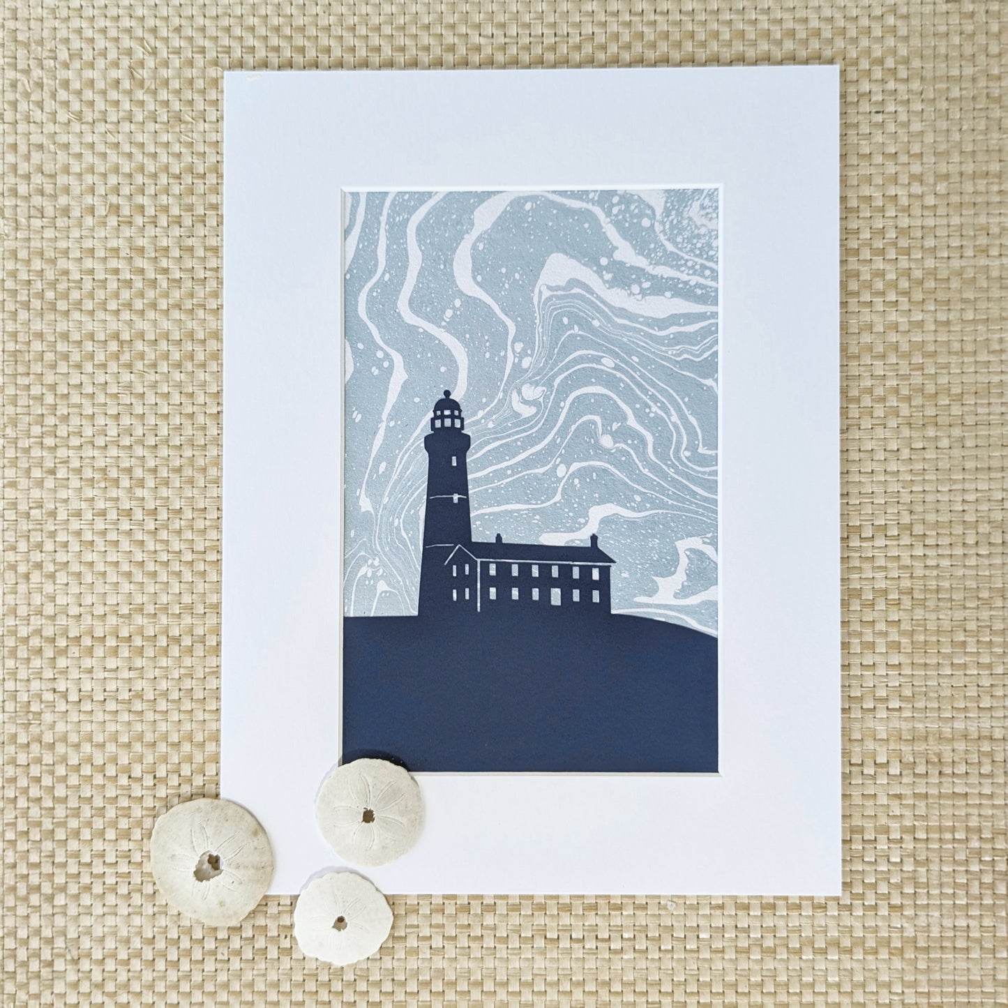 Montauk Lighthouse cut-paper silhouette over swirling marbled background, matted and styled with natural sand dollars on a natural woven surface.