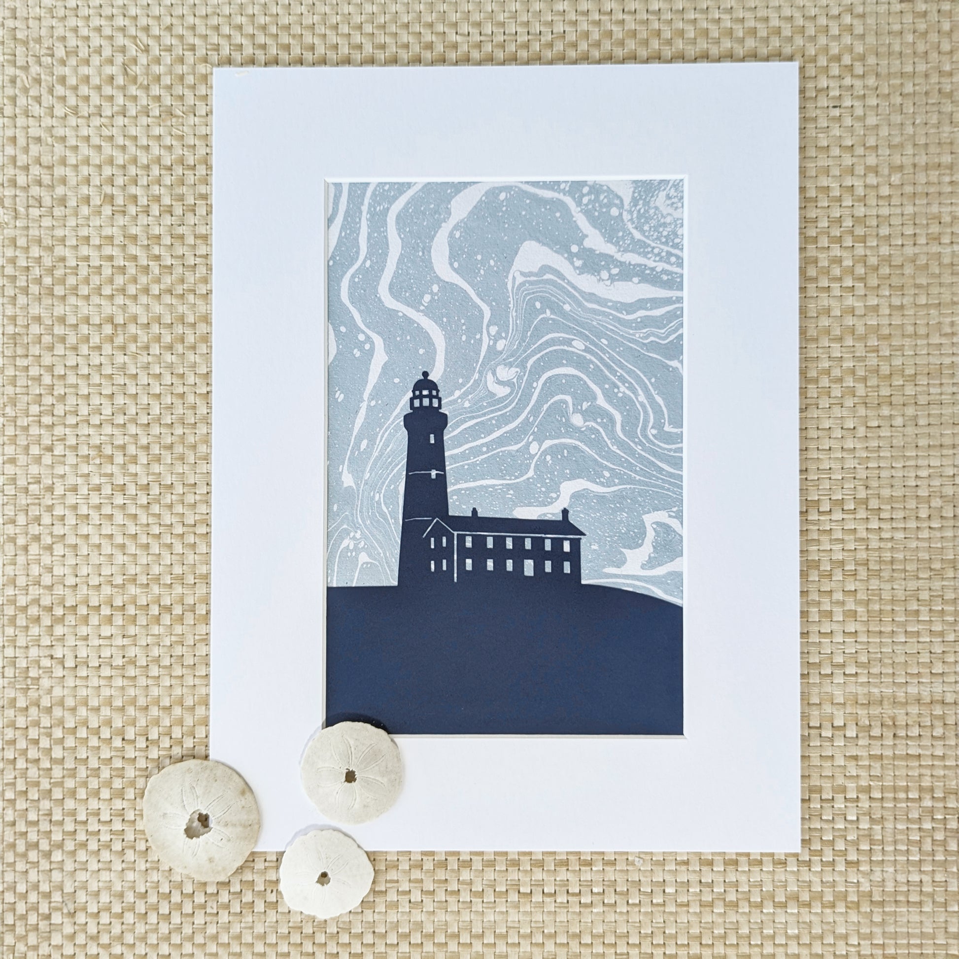 Montauk Lighthouse cut-paper silhouette over swirling marbled background, matted and styled with natural sand dollars on a natural woven surface.