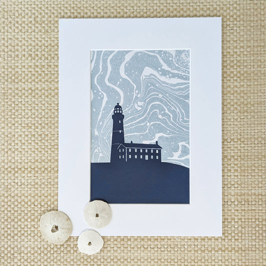 Montauk Lighthouse cut-paper silhouette over swirling marbled background, matted and styled with natural sand dollars on a natural woven surface.