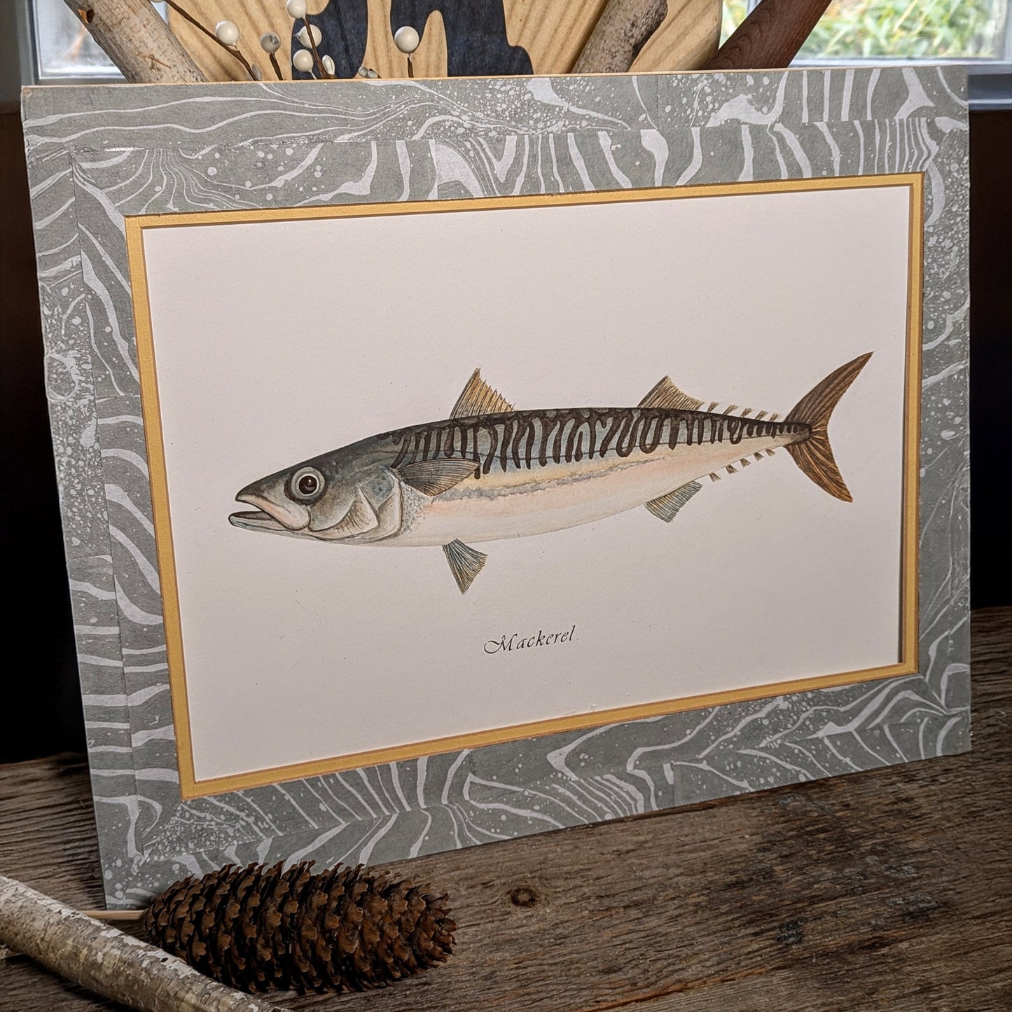 Double-matted Mackerel watercolor displayed on a rustic wood surface with gray suminagashi-marbled mat and coastal natural elements.