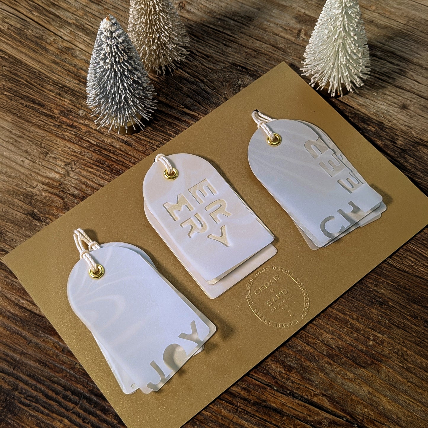 Three Suminagashi layered gift tag pairs—JOY, MERRY, and CHEER—displayed on a gold card with small bottlebrush trees.