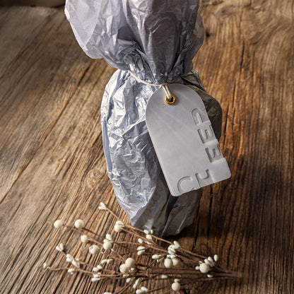 CHEER Suminagashi gift tag attached to a silver-wrapped bottle, styled on a rustic wood surface with winter berry stems.