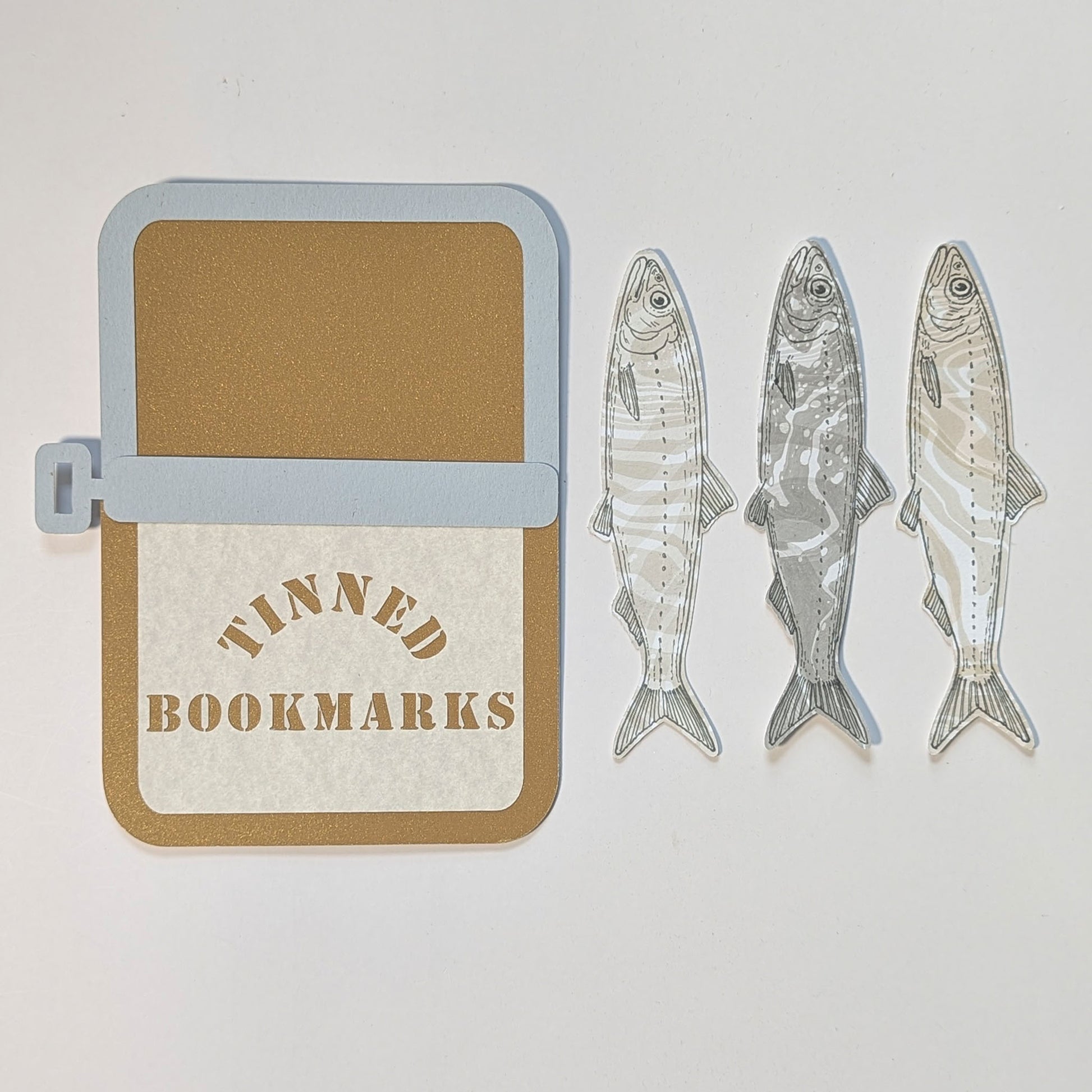 Custom die-cut paper sardine tin displayed beside three handmade sardine bookmarks with hand-drawn illustrations and marbled backs.