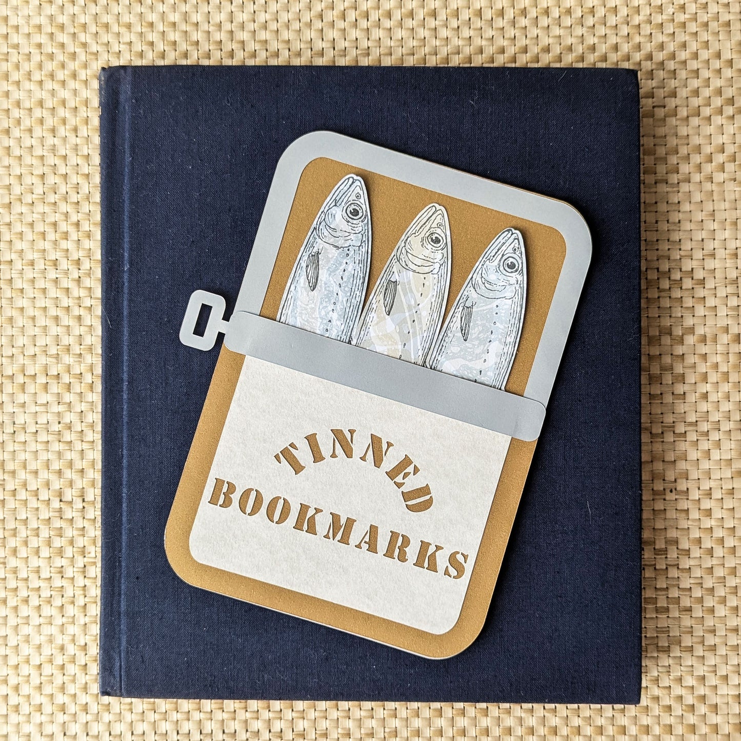 Handmade Tinned Sardines Bookmark Set resting on a navy linen hardcover book. Three hand-drawn sardine bookmarks with unique Suminagashi marbled patterns peek from a custom paper sardine tin by CEDAR Y SAND.