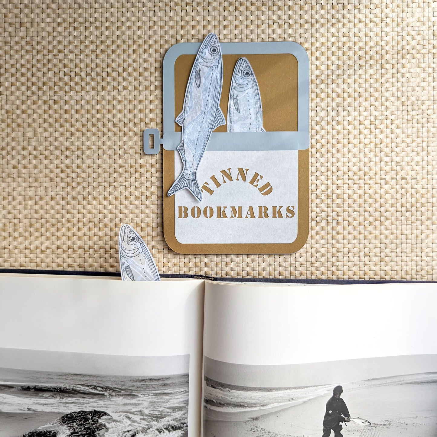Three handmade sardine bookmarks in a paper sardine tin with one bookmark tucked inside an open book showing the illustrated fish peeking from the page.