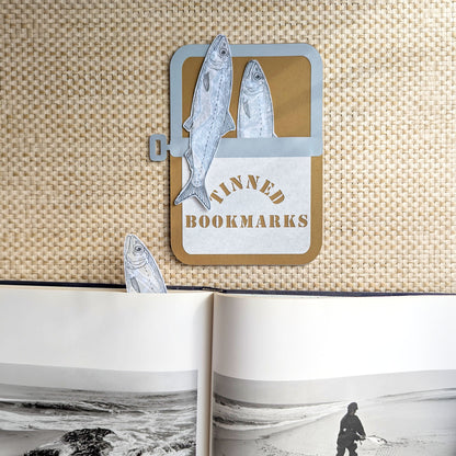Three handmade sardine bookmarks in a paper sardine tin with one bookmark tucked inside an open book showing the illustrated fish peeking from the page.