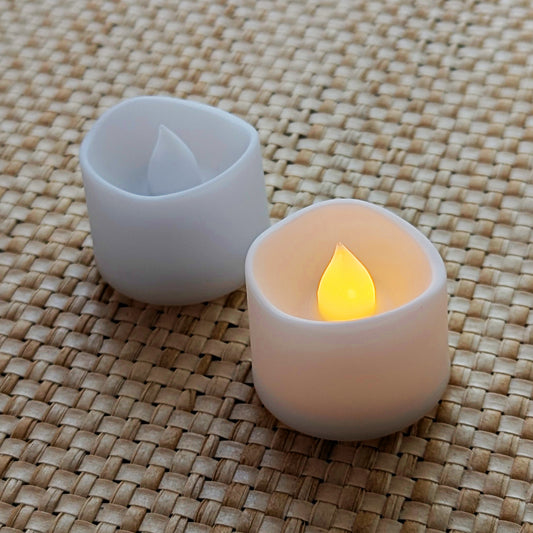 Two Autumn Glow Tealights, one lit and one unlit, showing gentle amber glow on textured woven background.