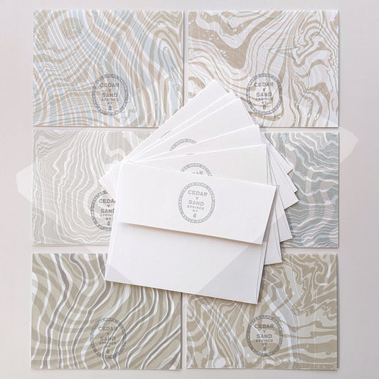 Back view of six Double Suminagashi notecards with Cedar Y Sand gray stamped insignia, shown with matching envelopes.