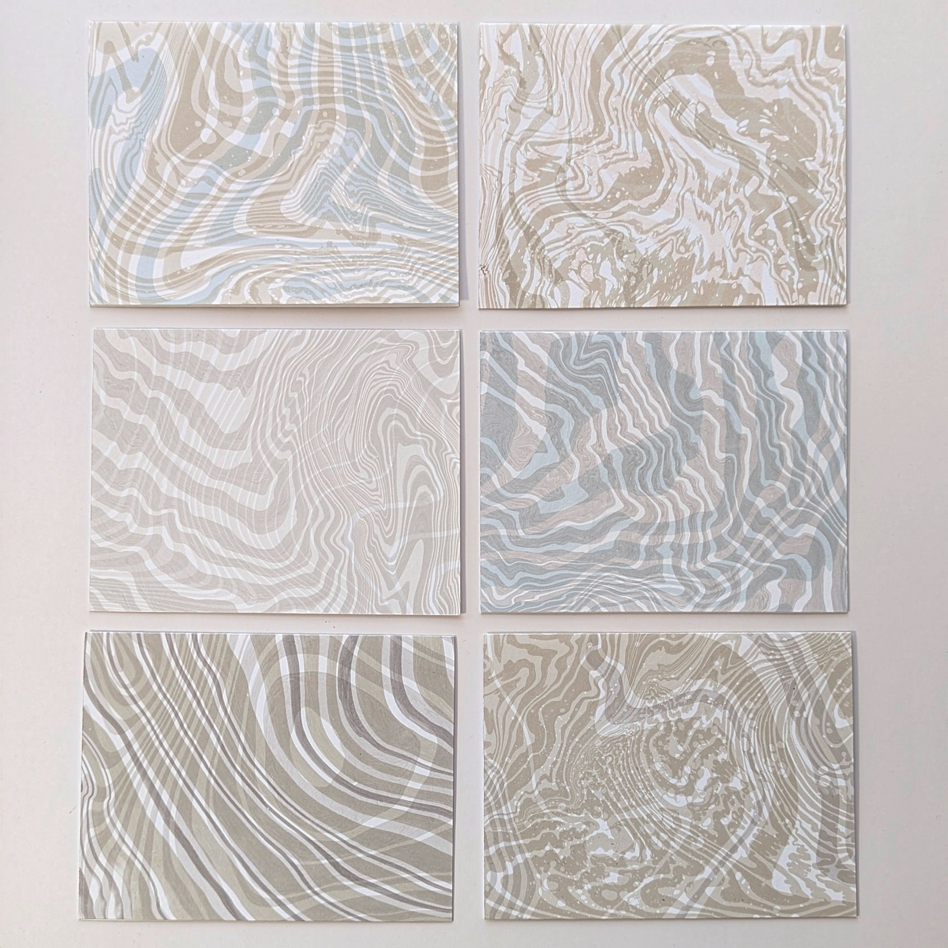 Frontside of set of six Double Suminagashi notecards laid flat, each with unique hand-marbled patterns in sandy beige, soft aqua, and pebble gray tones.