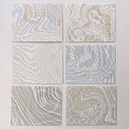 Frontside of set of six Double Suminagashi notecards laid flat, each with unique hand-marbled patterns in sandy beige, soft aqua, and pebble gray tones.