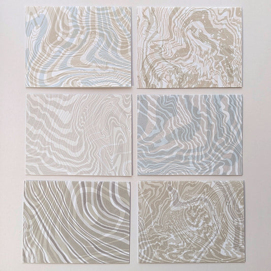 Frontside of set of six Double Suminagashi notecards laid flat, each with unique hand-marbled patterns in sandy beige, soft aqua, and pebble gray tones.