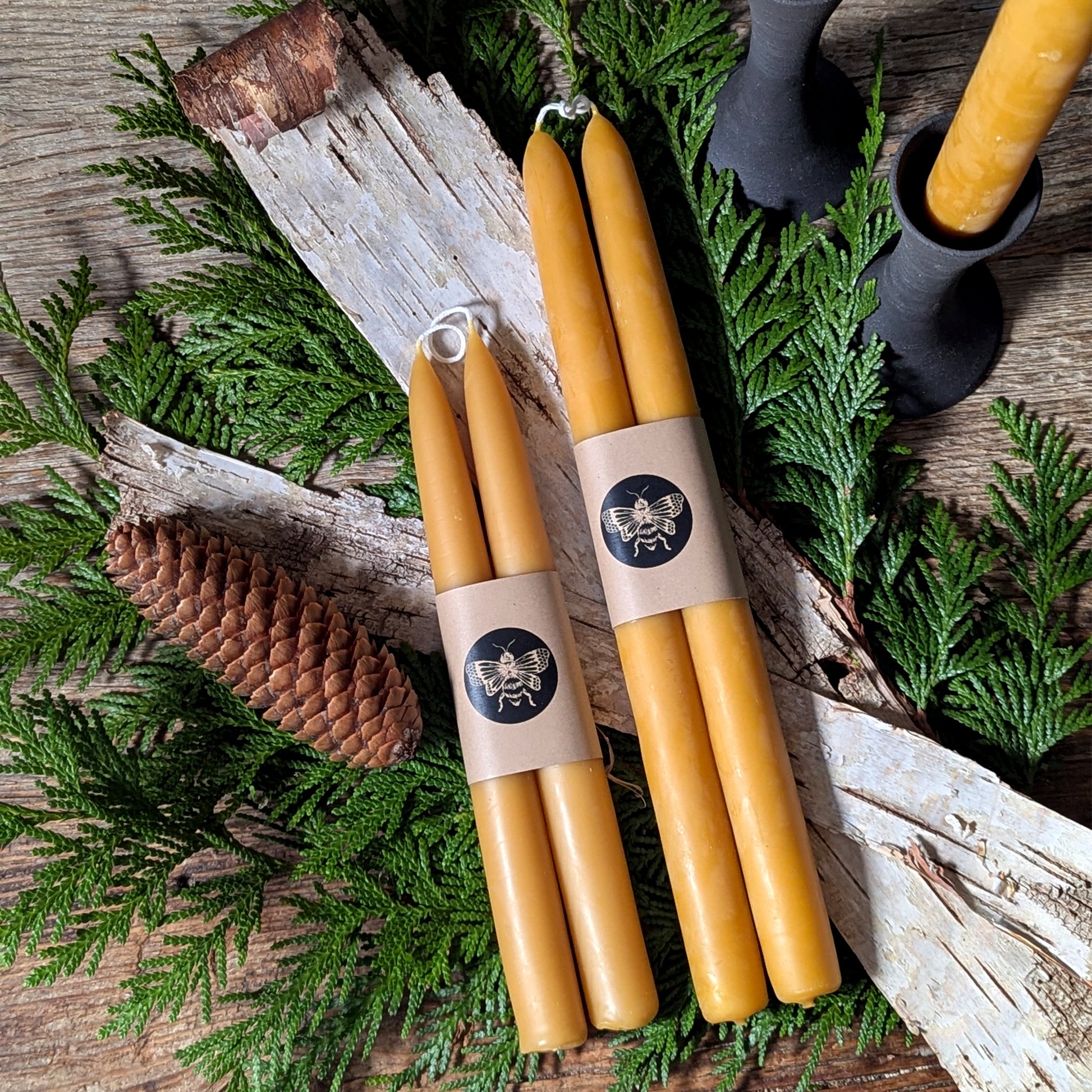 Pure beeswax candlesticks from Waxing & Raining displayed on a rustic wood table with cedar greenery, birch bark, and pinecones—natural, handcrafted home décor and candlelight styling.